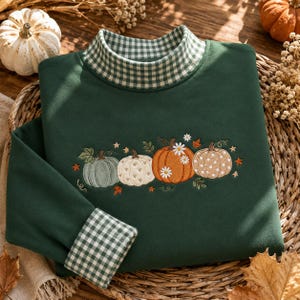 May include: A dark green long-sleeve sweatshirt featuring a mock neck collar and sleeve cuffs with a green and white gingham pattern. The chest displays an embroidered design of four pumpkins in shades of green, cream, and orange with daisies.