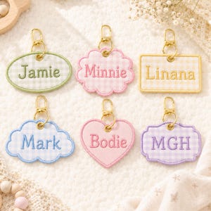 May include: Six fabric keychains with gold-tone lobster clasps. Shapes include an oval with 'Jamie', a scalloped circle with 'Minnie', a rectangle with 'Linana', a cloud with 'Mark', a heart with 'Bodie', and a plaque with 'MGH'.