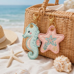 May include: Two embroidered keychains featuring a light mint green seahorse with a pink belly and a light pink starfish with a teal center design. Both items include gold-toned metal lobster claw clasps for attachment to bags or keys.