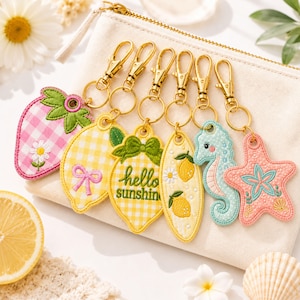 May include: Six embroidered keychains with gold-tone lobster clasps, featuring a pink gingham strawberry, a yellow gingham lemon with a bow, a lemon with the text hello sunshine, a surfboard with lemons, a light blue seahorse, and a pink starfish.