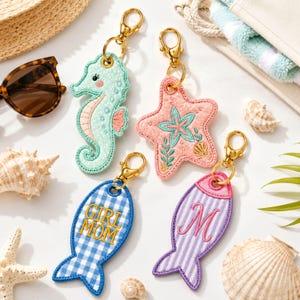 May include: Four embroidered keychains with gold-tone lobster clasps. One mint green seahorse, one pink starfish with a teal star design, one blue and white gingham fish with the text GIRL MOM, and one purple and white striped fish with a letter M.