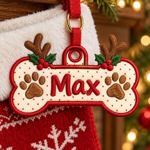May include: A bone-shaped embroidered ornament featuring the name Max in red thread. The cream-colored center has red polka dots and two brown paw prints. It is decorated with brown reindeer antlers, green holly leaves, and red berries.
