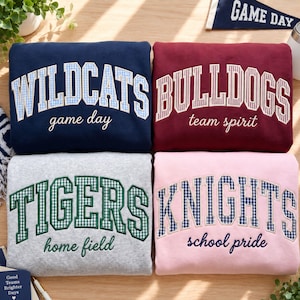 Może przedstawiać: Cztery złożone bluzy w kolorach granatowym, bordowym, szarym melanżowym i jasnoróżowym. Granatowa bluza ma napis WILDCATS game day, bordowa BULLDOGS team spirit, szara TIGERS home field, a różowa KNIGHTS school pride wykonane haftem.