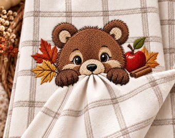 May include: A cream-coloured kitchen towel featuring a brown plaid pattern and an embroidered design of a brown bear peeking over a fold. The bear is surrounded by orange and yellow autumn leaves, a red apple with a green leaf, and two cinnamon sticks.