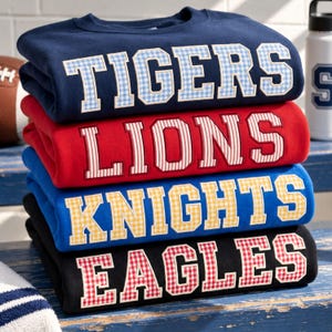 May include: A stack of four crewneck sweatshirts in navy, red, royal blue, and black. Each features large collegiate-style lettering with patterned fabric appliques: TIGERS in blue gingham, LIONS in red stripes, KNIGHTS in yellow gingham, and EAGLES in red gingham.