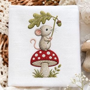 Peut inclure: Un carré de tissu blanc avec une broderie représentant une petite souris marron clair debout sur un champignon rouge à pois blancs. La souris tient une feuille de chêne verte et un petit gland. Des brins d'herbe verts sont brodés à la base.