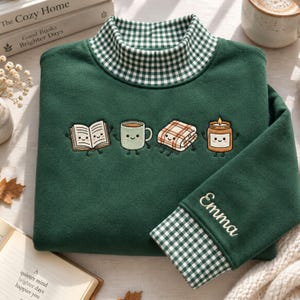 Puede incluir: Una sudadera verde oscuro con cuello alto de patrón de cuadros vichy verde y blanco. El pecho muestra personajes bordados de dibujos animados de un libro, una taza, una manta y una vela. El puño de la manga tiene el nombre Emma bordado.