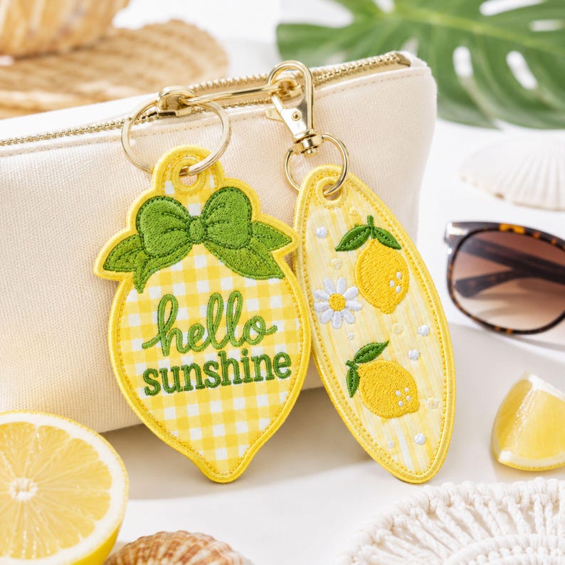May include: Two embroidered keychains attached to a gold-tone metal ring. One keychain is lemon-shaped with a yellow gingham pattern, a green bow, and the text hello sunshine. The other is an oval shape with yellow lemons and a white flower.