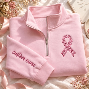 May include: A light pink quarter-zip sweatshirt featuring a silver metal zip and a high collar. The chest displays an embroidered awareness ribbon composed of small pink hearts, while the sleeve is embroidered with the text custom name.
