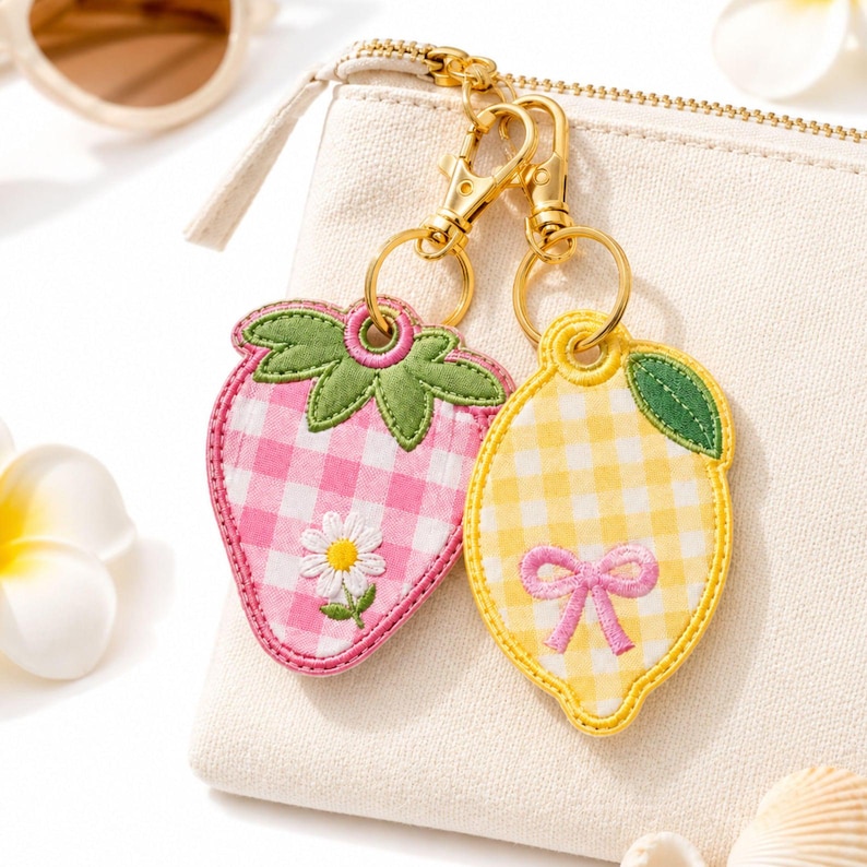 May include: Two embroidered keychains attached to a gold-toned metal clasp. One keychain is a pink and white gingham strawberry with a green stem and a white daisy. The other is a yellow and white gingham lemon with a green leaf and a pink bow.