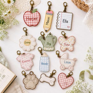 May include: A collection of twelve embroidered fabric keychains with bronze-toned lobster clasps. Shapes include an apple, pencil, notebook, flower, watering can, butterfly, onesie, baby bottle, stroller, paw print, bone, and heart, each featuring a name.