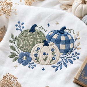May include: A white fabric towel featuring an embroidered design of three pumpkins. One pumpkin is sage green with white leaf patterns, one is cream with blue floral details, and one features a blue and white checkered pattern with blue stems.