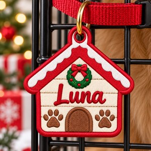 Könnte beinhalten: Ein hausförmiges Haustier-Namensschild mit roter und cremefarbener Stickerei. Das Design zeigt einen grünen Kranz mit roter Schleife, den Namen Luna in roter Schreibschrift, eine braune Bogentür und zwei braune Pfotenabdrücke.
