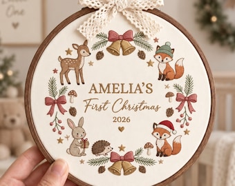 May include: A circular wooden embroidery hoop containing white fabric with a decorative wreath design. The embroidery features a deer, two foxes, a rabbit, and a hedgehog, with text reading AMELIA'S First Christmas 2026. A cream lace bow is attached to the top.