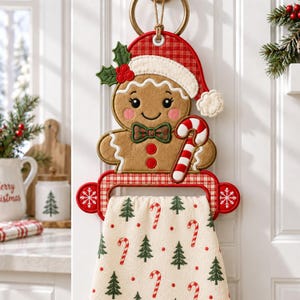 May include: A hanging kitchen towel holder featuring a plush gingerbread person wearing a red and white chequered Santa hat, a green bow tie, and holding a candy cane. The attached cream-coloured towel is decorated with a pattern of green pine trees and red candy canes.