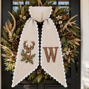 Peut inclure: Une couronne décorative composée de branches de pin vertes, de pommes de pin et de bois de cerf. Un ruban en tissu rayé beige et blanc à bords festonnés est suspendu au centre, orné d'une tête de cerf brodée et de la lettre W en fil marron.