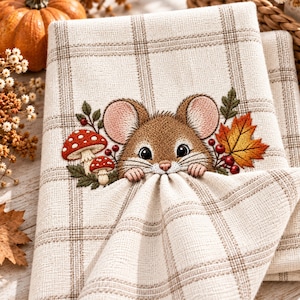 May include: A beige and brown plaid fabric featuring an embroidered brown mouse peeking over a fold. The embroidery includes two red and white spotted mushrooms, green leaves, red berries, and a single orange and yellow maple leaf accent.
