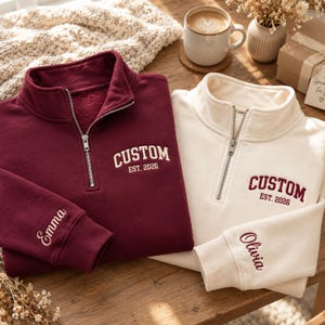 May include: Two quarter-zip sweatshirts, one in maroon and one in cream. The maroon sweatshirt features white embroidery reading "CUSTOM EST. 2026" on the chest and "Emma" on the sleeve. The cream sweatshirt has maroon embroidery reading "CUSTOM EST. 2026" and "Olivia".