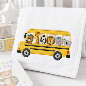 May include: A white blanket featuring an embroidered yellow school bus with a bear, giraffe, zebra, lion, elephant, and koala as passengers. The bus has black wheels and windows, and the blanket has a subtle white checkered border trim.