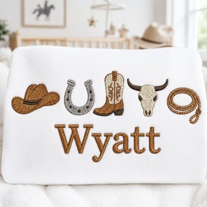May include: A white sweatshirt featuring embroidered western-themed icons including a brown cowboy hat, a grey horseshoe, a brown cowboy boot, a white steer skull, and a brown lasso. The name Wyatt is embroidered in brown below the icons.