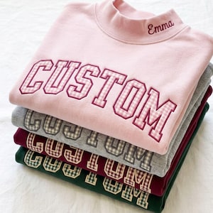 May include: A stack of four mock neck sweatshirts in light pink, gray, maroon, and dark green. Each sweatshirt features the word CUSTOM in varsity-style letters with a gingham pattern and the name Emma embroidered on the collar of the top garment.