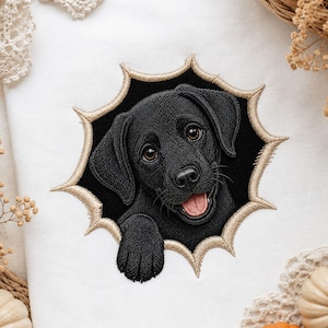 May include: An embroidered patch on white fabric featuring a black Labrador puppy peeking through a jagged, star-shaped black hole with a tan stitched border. The puppy has brown eyes, a pink tongue, and one paw visible in the foreground.