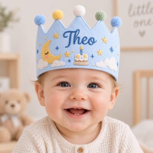 Könnte beinhalten: Eine hellblaue Stoffkrone mit dem gestickten Namen Theo in dunkelblauer Schreibschrift. Die Krone ist mit einem gelben Halbmond, weißen Wolken, gelben Sternen und einer kleinen Geburtstagstorte mit Kerze verziert und hat fünf Bommeln oben.