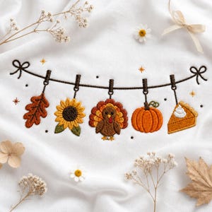 May include: A white fabric featuring an embroidered clothesline design with five hanging items: an orange oak leaf, a yellow sunflower, a brown turkey, an orange pumpkin, and a slice of pumpkin pie with white whipped cream on top.