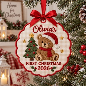 Puede incluir: Un adorno navideño de tela color crema con borde festoneado y lazo rojo. Presenta un oso de peluche marrón bordado con gorro y bufanda de Papá Noel rojos, un árbol de Navidad verde y el texto Olivia's FIRST CHRISTMAS 2026.