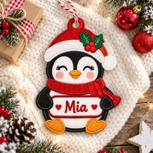 Può includere: Una decorazione natalizia in feltro a forma di pinguino che indossa un cappello da Babbo Natale rosso con foglie di agrifoglio verdi e bacche rosse. Il pinguino ha una sciarpa rossa, piedi arancioni e un cartello bianco con il nome Mia e due cuori.