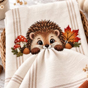 May include: A cream-coloured kitchen towel with vertical tan stripes featuring an embroidered design of a brown hedgehog, two red and white spotted mushrooms, a brown acorn, and a vibrant orange and red maple leaf on the fabric surface.
