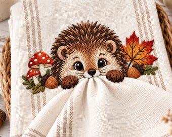 May include: A cream-coloured kitchen towel with vertical tan stripes featuring an embroidered design of a brown hedgehog, two red and white spotted mushrooms, a brown acorn, and a vibrant orange and red maple leaf on the fabric surface.