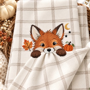 May include: A cream-colored kitchen towel featuring a brown plaid pattern and an embroidered fox face with dark brown paws. The design includes a single orange maple leaf, a small orange pumpkin, a yellow crescent moon, and two black stars.