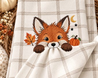 May include: A cream-coloured kitchen towel featuring a brown plaid pattern and an embroidered fox face with dark brown paws. The design includes a single orange maple leaf, a small orange pumpkin, a yellow crescent moon, and two black stars.