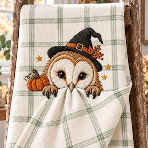 May include: A cream-coloured kitchen towel featuring a green plaid pattern and an embroidered design of a brown owl wearing a black witch hat. The owl is positioned next to an orange pumpkin, with small yellow stars surrounding the figure.