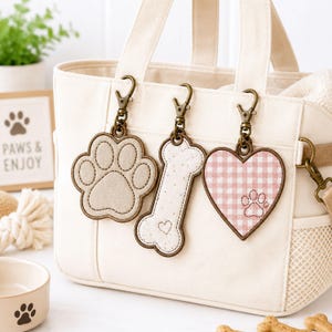 Puede incluir: Tres dijes colgantes sujetos a un bolso de lona color crema. Los dijes incluyen una huella de pata beige, un hueso blanco moteado con un corazón pequeño y un corazón de cuadros vichy rosa y blanco con una huella pequeña, con broches bronce.