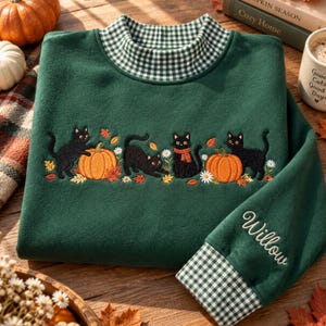 Op de afbeelding: Een donkergroen sweatshirt met een opstaande kraag in een wit en groen vichy-ruitpatroon. De voorzijde toont een borduursel van drie zwarte katten, oranje pompoenen, herfstbladeren en witte madeliefjes. De naam Willow is op de mouw geborduurd.