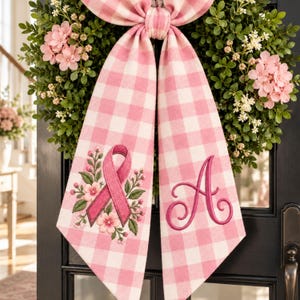 Op de afbeelding: Een decoratieve krans met groen blad en roze bloemen, versierd met een grote roze en wit geblokte strik. Eén lintuiteinde toont een geborduurd roze bewustzijnslint omringd door bloemen, terwijl de andere een grote letter A bevat.