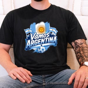 May include: A black t-shirt featuring a graphic print with the text Vamos ARGENTINA in white and blue lettering. The design includes a central sun emblem, two small Argentine flags, and a soccer ball at the bottom against a blue brushstroke background.