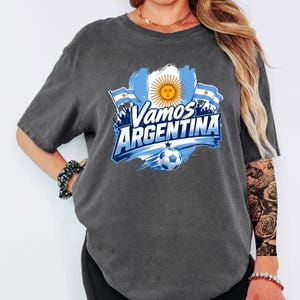 May include: A charcoal gray short-sleeve t-shirt featuring a graphic print with the text Vamos ARGENTINA, the Argentine flag, a golden sun emblem, and a soccer ball. The design includes blue and white brushstroke accents and silhouettes.