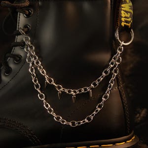 May include: A black leather ankle boot featuring a double-layered silver-toned metal chain accessory. The chain is attached to the boot's eyelets and heel loop, and it is decorated with small, pointed silver spikes hanging from the lower chain.