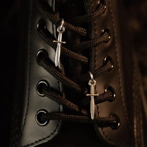 May include: Two small silver-toned sword charms attached to the black laces of a black leather boot. The charms are shaped like miniature daggers with a pointed blade and a crossguard, hanging securely from the woven laces between the metal eyelets.