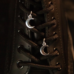 May include: Two silver-toned crescent moon charms attached to the black laces of a black boot. The charms are small, metallic, and feature a smooth, curved design. They are positioned on the crisscrossed laces, adding a decorative element.