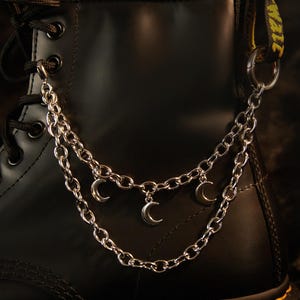 May include: A black leather ankle boot featuring a double-layered silver-toned metal chain accessory. The chain is attached to the eyelets and the heel loop, adorned with three small silver crescent moon charms hanging from the lower chain link.