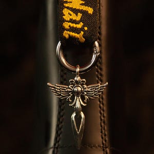 May include: A silver-toned metal charm featuring a winged design with a central sphere and a pointed base, attached to a circular ring. The charm is suspended from a black fabric pull tab with yellow embroidered text that reads AirWair.