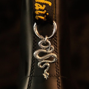 Op de afbeelding: Een zilverkleurige slangenbedel bevestigd aan een ronde metalen ring, die door een zwarte stoffen hiellus met geel geborduurde tekst Dr. Martens is gehaald, tegen een glad oppervlak van zwart leer.
