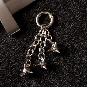 May include: A silver-toned metal charm featuring a circular ring attached to three short chain links, each ending in a small, four-pointed star-shaped spike. The metallic finish is reflective, and the design is symmetrical with dangling elements.