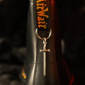 May include: A black boot featuring a heel pull tab with yellow text that reads AirWair. A silver-toned metal sword charm hangs from a circular ring attached to the pull tab. The boot has a smooth, dark surface with visible vertical stitching.