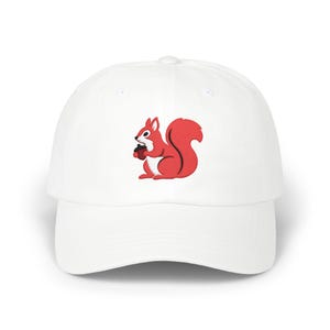 Könnte beinhalten: Eine weiße Baseballkappe mit einer Stickerei eines roten Eichhörnchens, das eine kleine braune Eichel hält. Das Eichhörnchen hat eine weiße Brust und schwarze Umrisse und ist mittig auf der Vorderseite platziert. Die Kappe hat einen gebogenen Schirm.