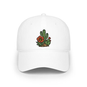 Könnte beinhalten: Eine weiße Baseballkappe mit einer mittigen Stickerei, die einen grünen Saguaro-Kaktus, einen kleineren Feigenkaktus und eine große orangefarbene Blüte mit gelber Mitte zeigt, die auf braunen Steinen mit grünen Blättern am Fuß ruhen.